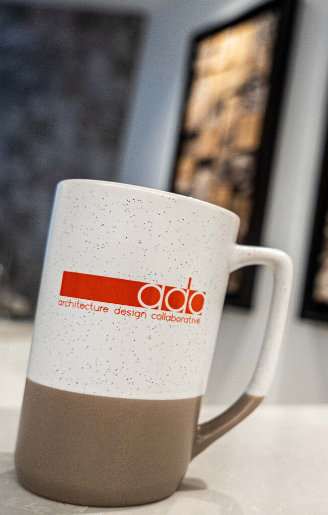 A ceramic mug with a speckled white upper half and solid beige lower half, featuring the red text and logo "architecture design collaborative." Two blurred pictures are in the background.