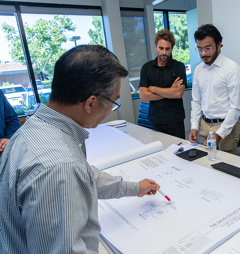 Three men are standing around a table covered with commercial architecture blueprints. One man is pointing at the designs with a pen, while the other two listen attentively to discuss their design services.