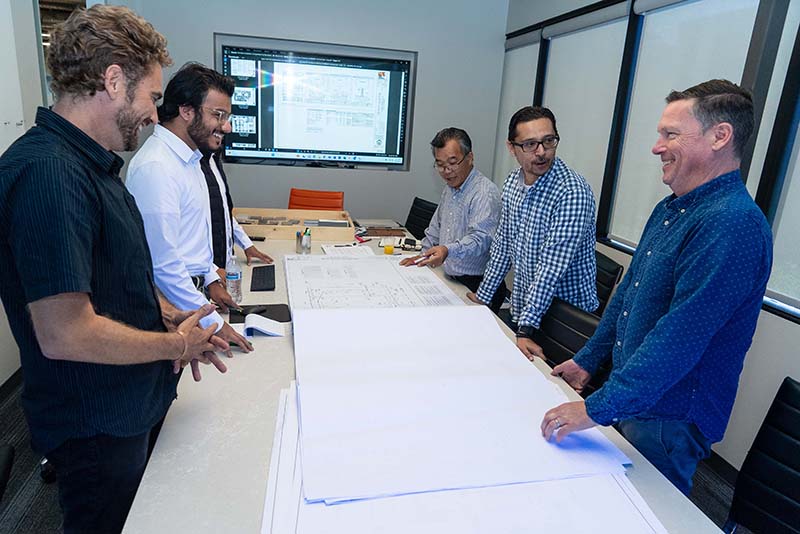Five men stand around a conference table reviewing large architectural blueprints, with a digital screen in the background displaying schematics. The scene epitomizes their commercial architecture design services in action, showcasing their collaborative approach and attention to detail.