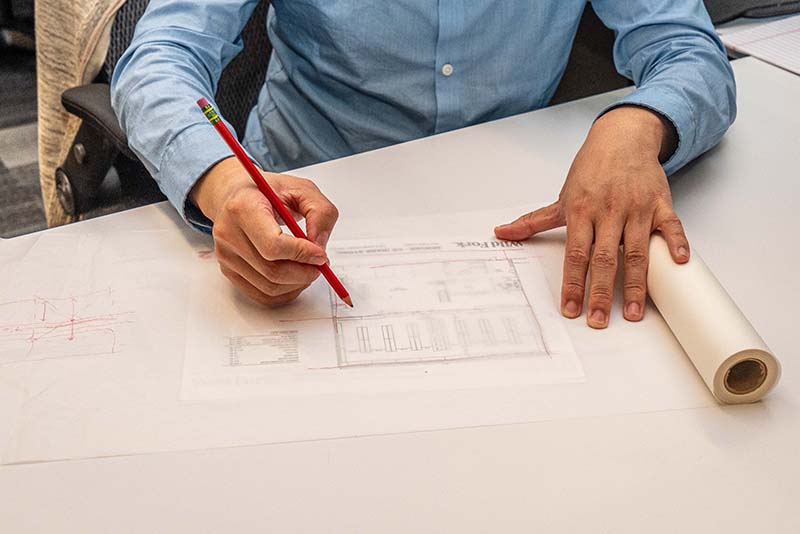 An entry level architect in a blue shirt is meticulously drawing architectural plans with a red pencil on a large sheet of paper, surrounded by additional sheets and a roll of paper on the table.
