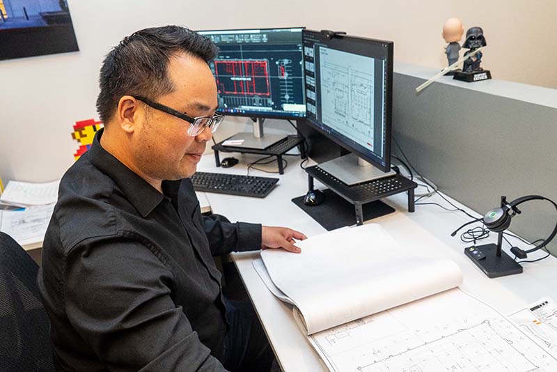 A man wearing glasses works at a desk with two computer monitors displaying technical drawings. He is looking at a large book or document with similar drawings in a modern office setting, immersed in his architecture job.