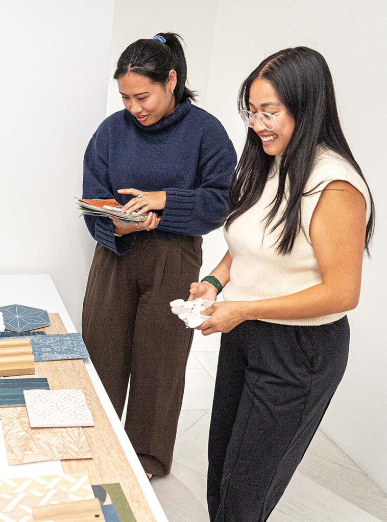 Two people stand by a table, examining various tile samples and design materials. Embracing the spirit of collaborative architecture design, one holds a set of tiles while the other smiles and looks at a tile-shaped object in their hands.