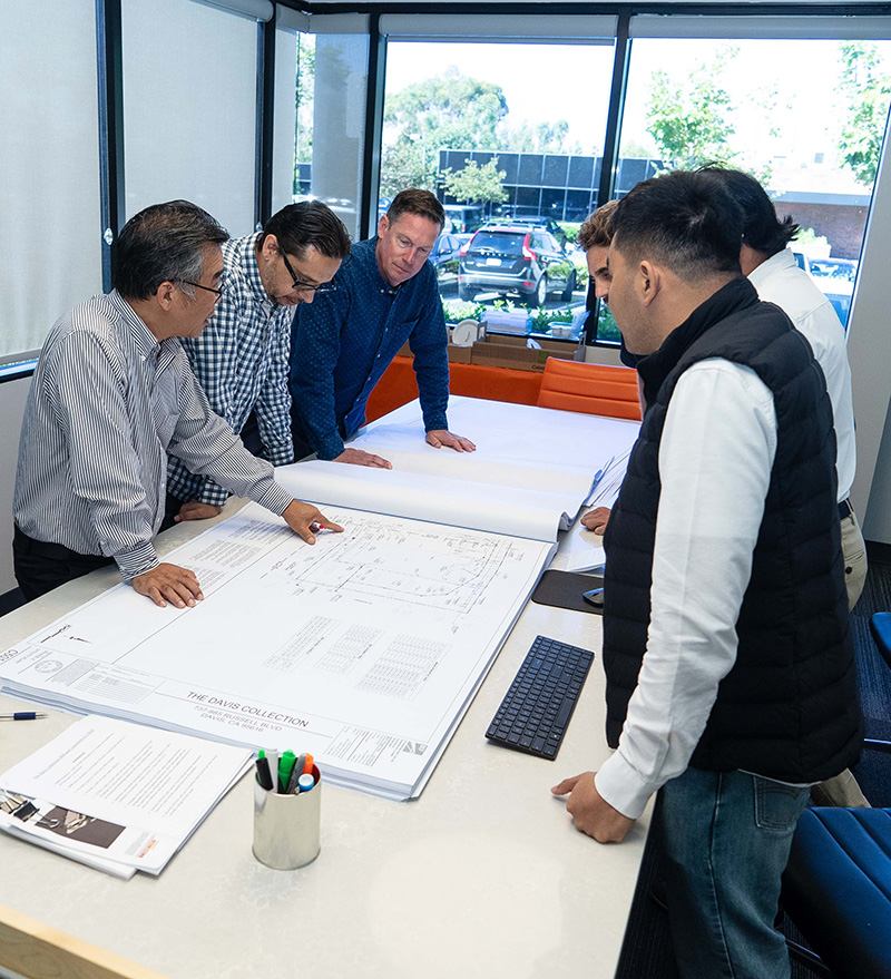 A group of five men are gathered around a table reviewing large architectural plans in a bright office space with windows, exemplifying the collaborative nature of architecture jobs.