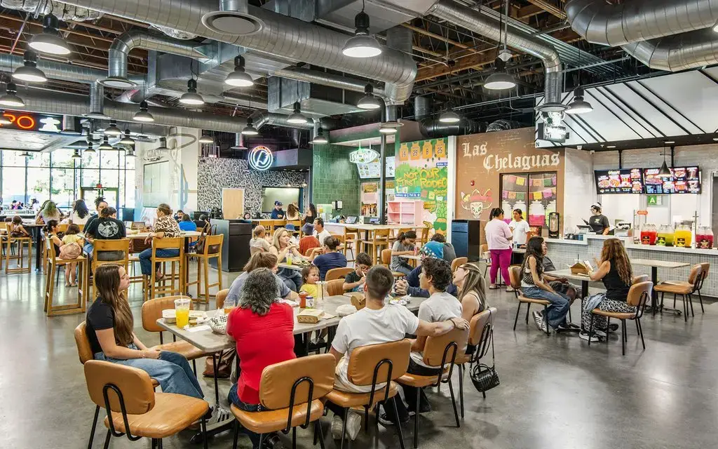 Rodeo 72 food hall in Whittier, California, with communal dining tables, tenant storefronts including Las Chelaguas, and an exposed industrial ceiling