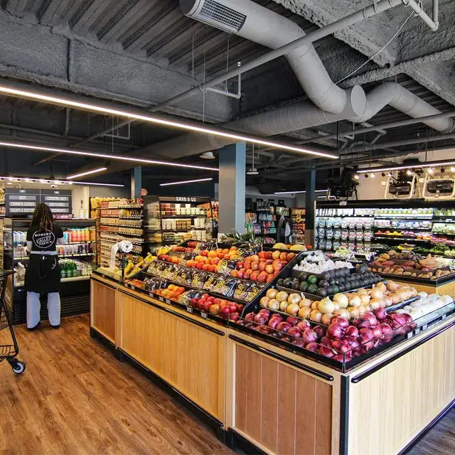 Grocery store interior with a central wood produce display of fruit and vegetables, exposed ceiling systems, and a shopper in an aisle