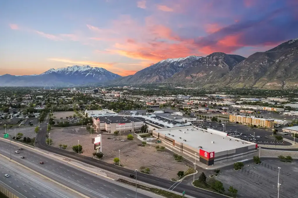 Aerial sunset view of a suburban shopping center beside a highway, with a Target store in the foreground, large parking lots, and snow-capped mountains under a pink and orange sky.