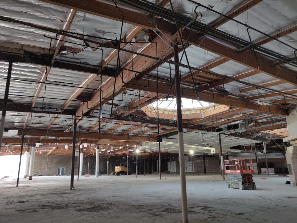 Interior of the former big-box store during renovation. Exposed wood roof beams, foil-faced insulation, and hanging conduit span an empty concrete floor, with a scissor lift and excavator in the open space.