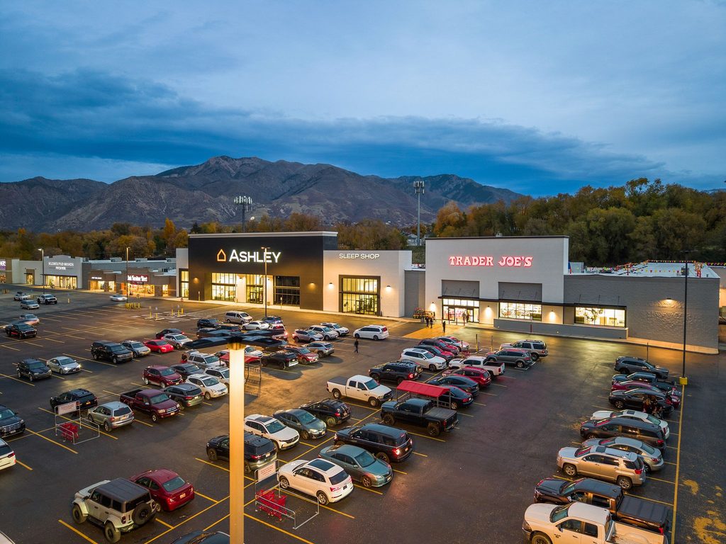 Twilight aerial view of the remodeled shopping center with Ashley Furniture, a Sleep Shop, and Trader Joe’s. Lit storefronts face a full parking lot, with mountains and a cloudy blue evening sky behind the buildings.