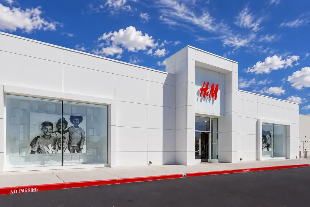 White H&M storefront with a red logo over the entrance, large window displays, and a red-painted curb under a bright blue sky.