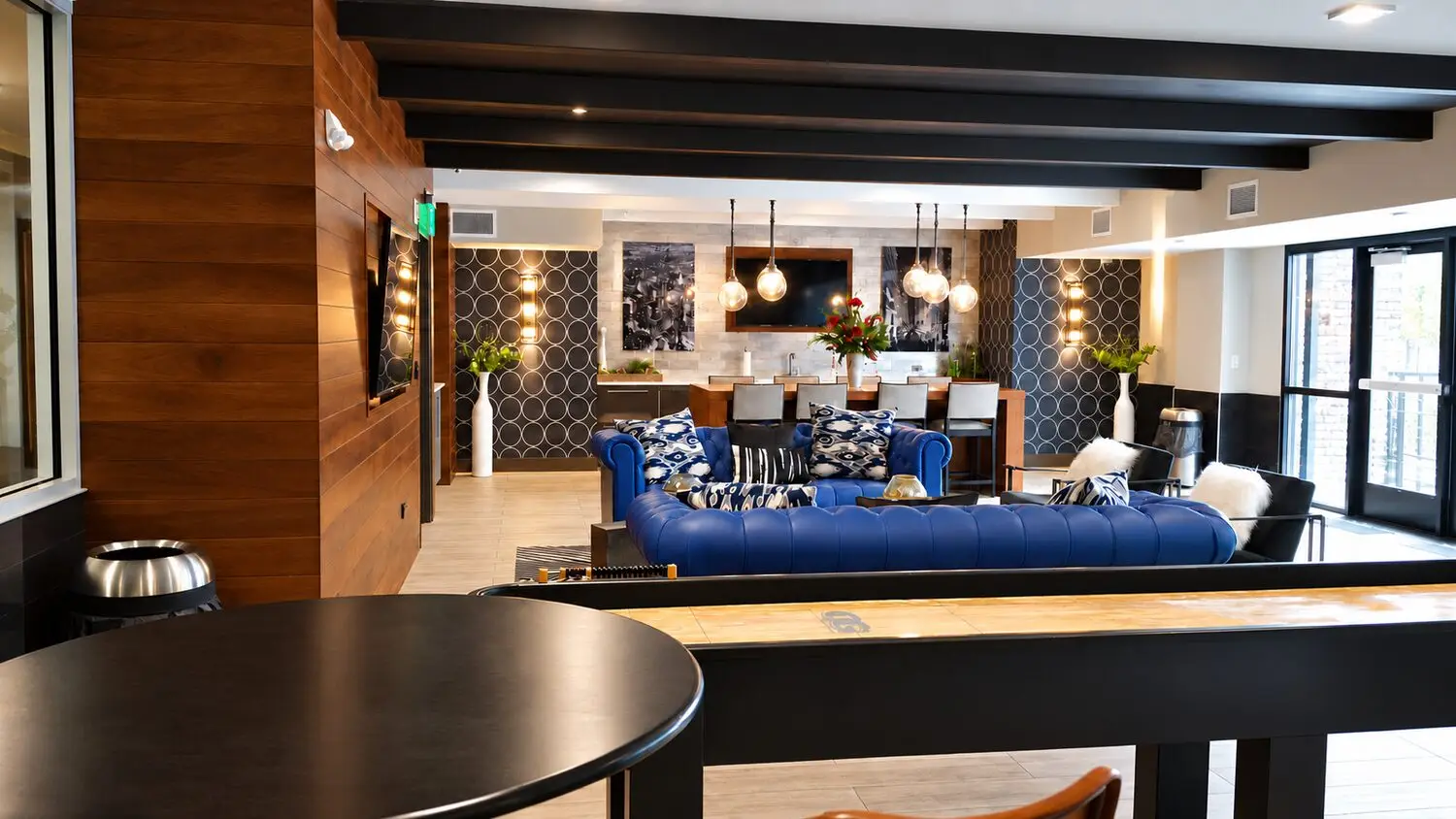 Amenity room looking past a shuffleboard table to a blue chesterfield and a wood bar with globe pendants. Circle-patterned tile, black beams, and patio doors frame the space.