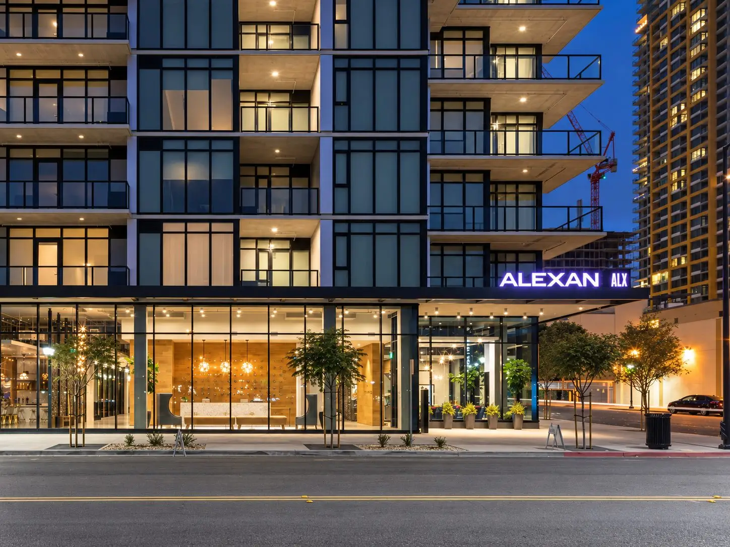Nighttime street-level view of the Alexan ALX apartment building in San Diego. The modern mid-rise has stacked glass balconies and a glowing ground-floor lobby behind floor-to-ceiling windows. An illuminated “ALEXAN ALX” sign sits above the entrance; a wood-paneled reception wall and hanging lights are visible inside. Young trees line the sidewalk.