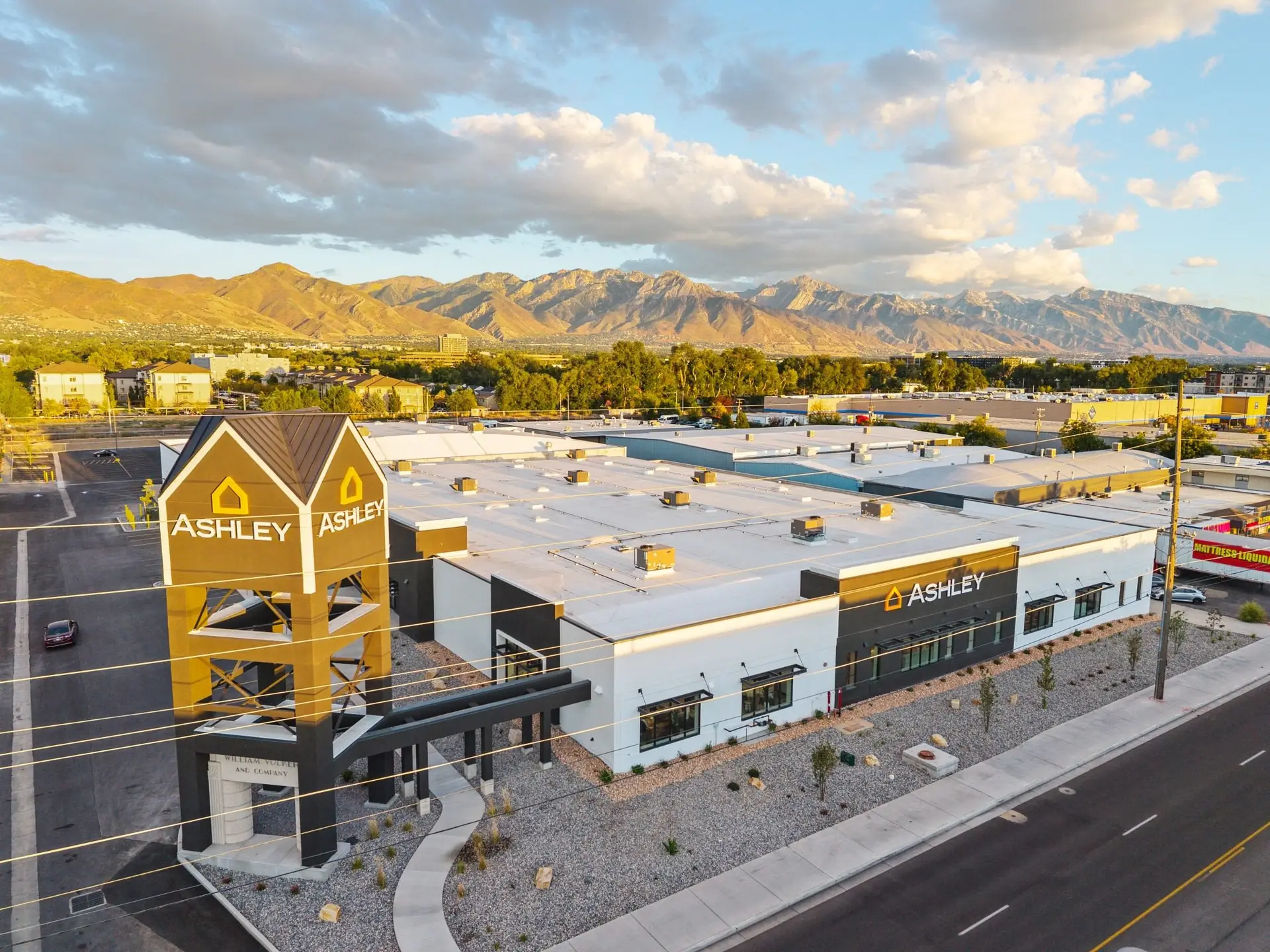 Aerial of Ashley Furniture Salt Lake City at dusk, with the branded tower over the historic William Volker entry and mountains beyond