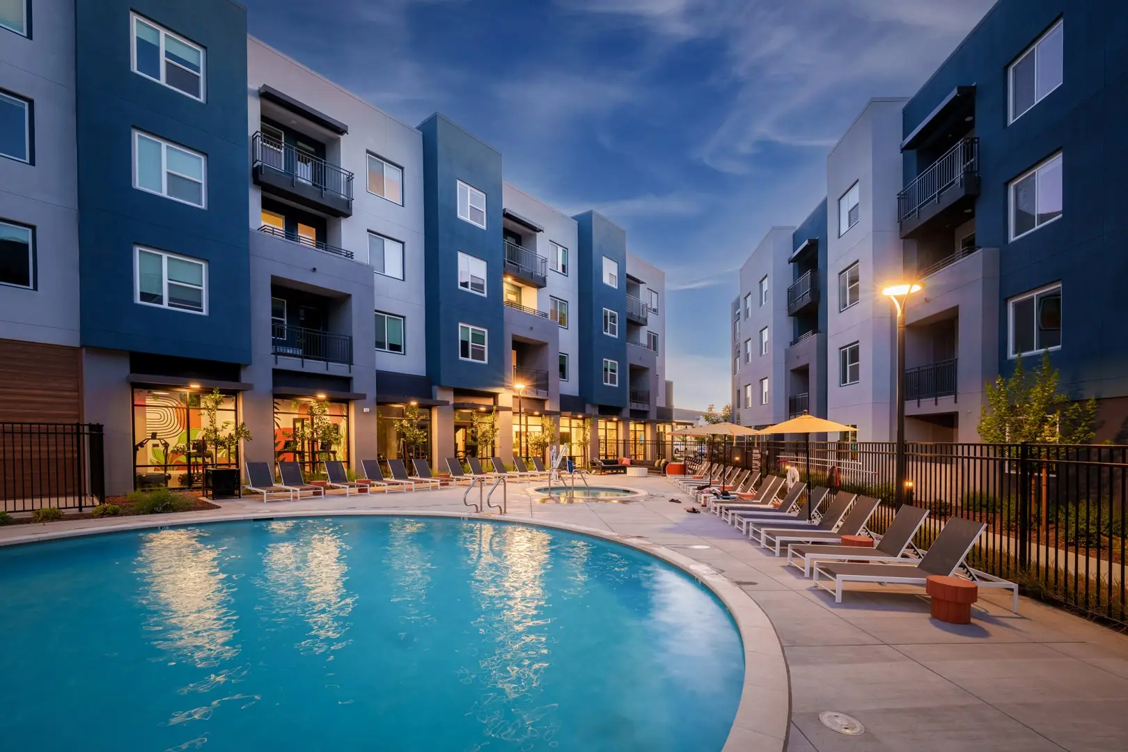 Courtyard pool at dusk, ringed by teal and gray apartment wings. A curved pool reflects building lights; lounge chairs line both sides, with umbrellas and a spa beyond a black fence. Lit windows and balconies face the deck.
