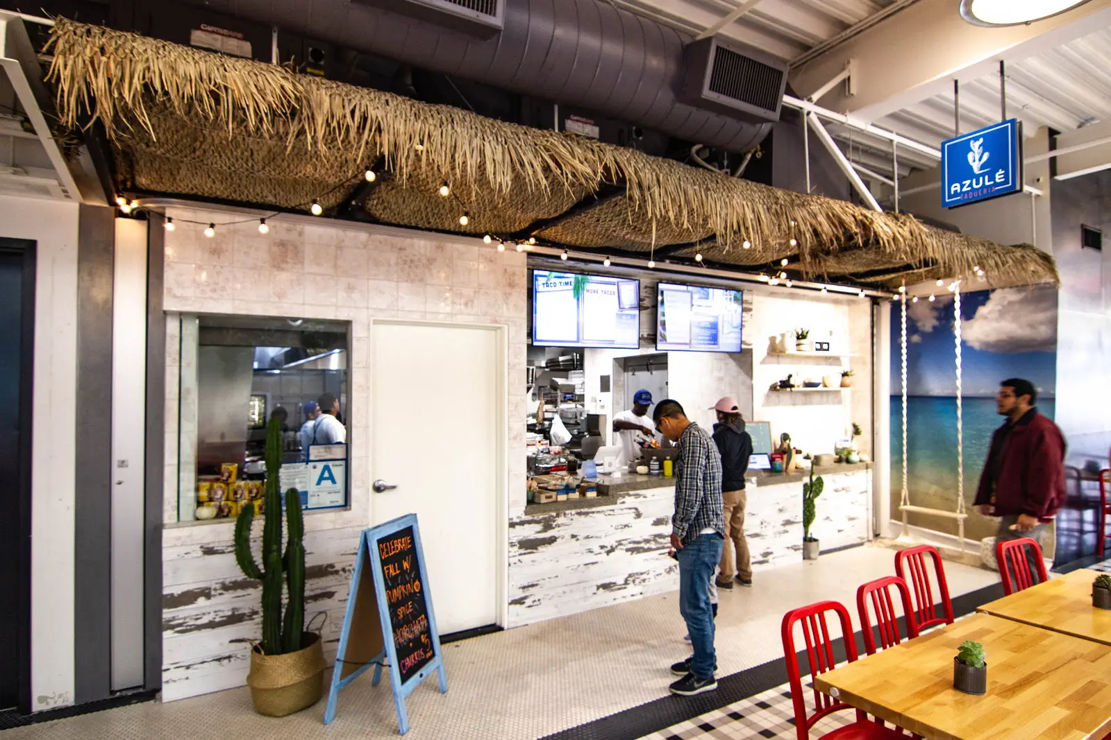 Azulé Taqueria stall in Santa Monica, California, with a thatched canopy and string lights, whitewashed wood counter, digital menus, a County health-grade A window, potted cactus, and an ocean mural with a rope swing.