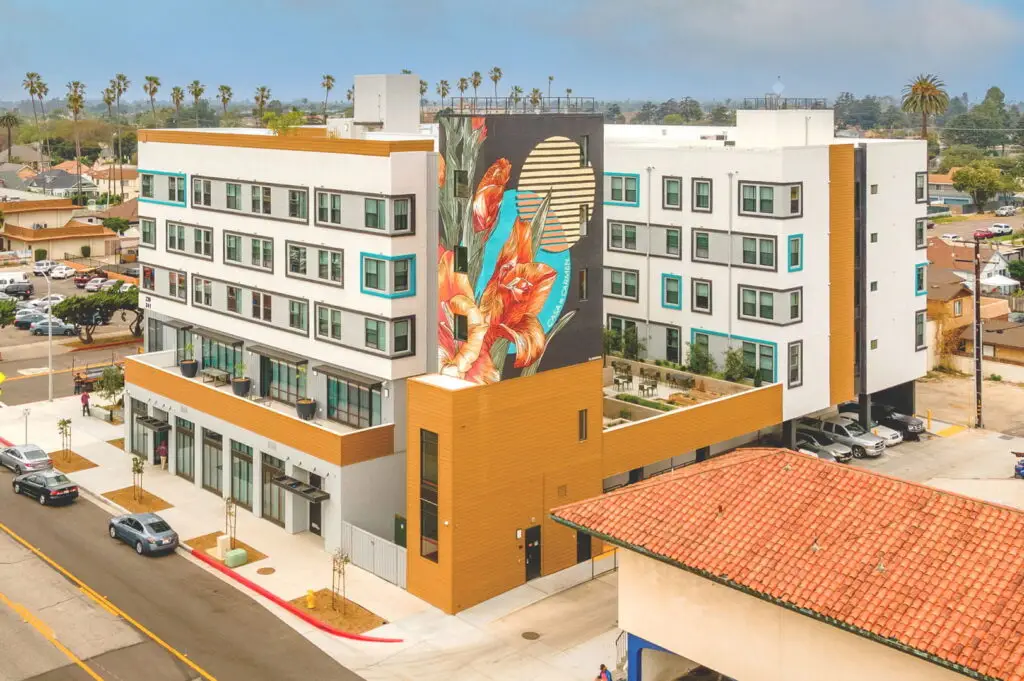 Aerial view of Casa de Carmen in Oxnard, a mid-rise apartment building in white and gray with mustard-gold panels and teal window accents. A tall mural of orange gladiolus and a striped sun disk reads “CASA DE CARMEN.” Street-level storefronts face the sidewalk; a raised courtyard and tuck-under parking sit behind. A red-tile roof is in the foreground; palms and neighborhood houses stretch beyond.