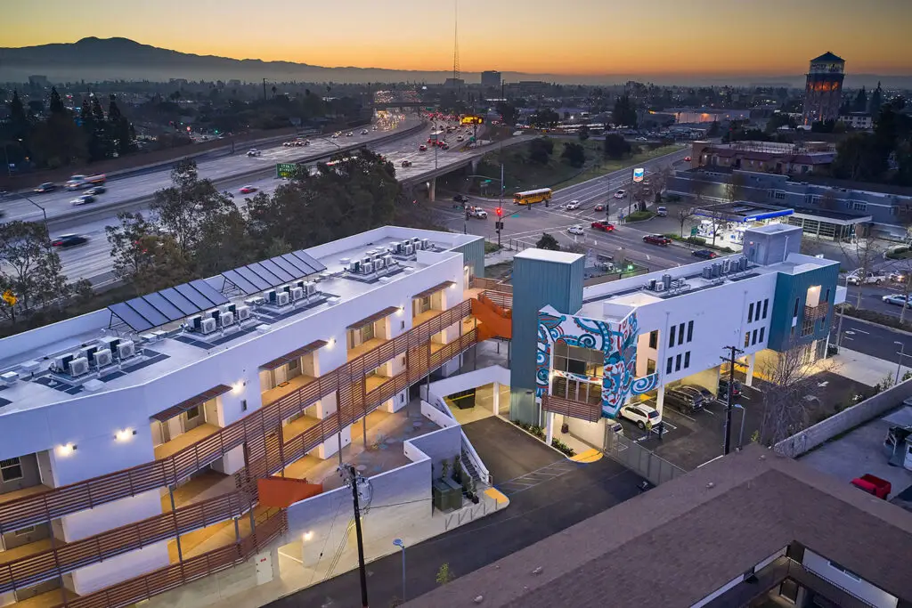 Aerial dusk view of Casa Querencia in Santa Ana, a white and teal multifamily building with wood-slat walkways, rooftop solar panels and HVAC units. A colorful mural wraps one corner. A freeway and glowing city sit beyond, with mountains on the horizon.
