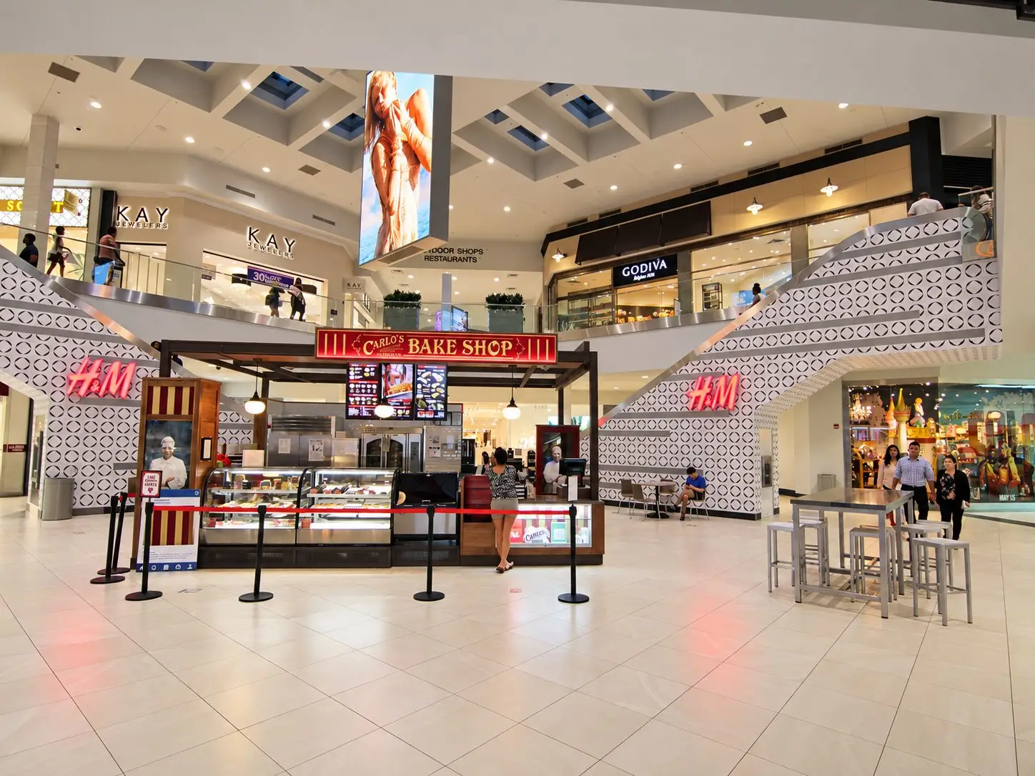 Two-level mall atrium with a Carlo’s Bake Shop kiosk in the center. A large vertical LED screen hangs from the ceiling. Patterned stair towers with H&M signs rise on both sides; Kay Jewelers and Godiva occupy the upper level. Shoppers walk the tiled floor.