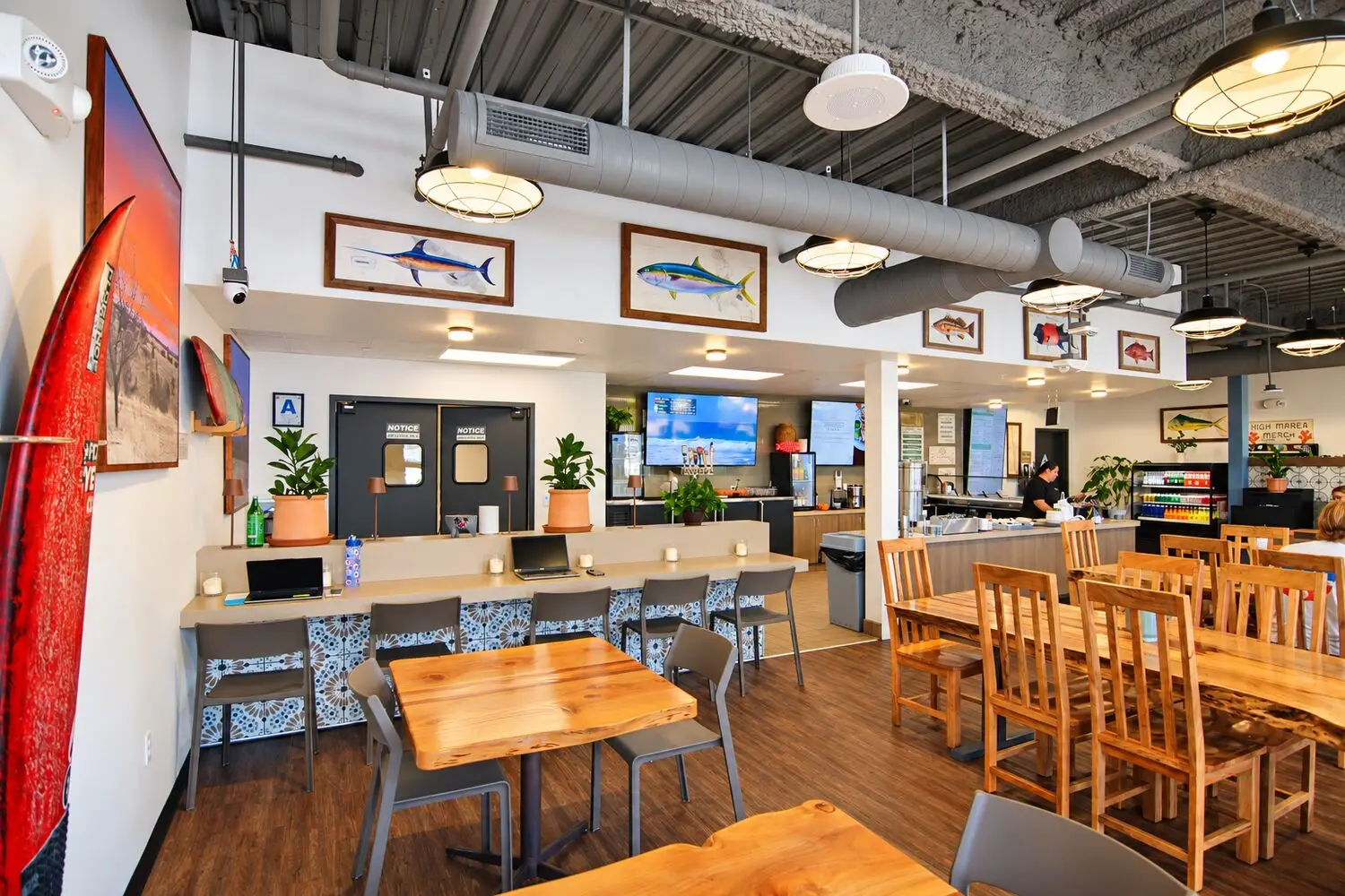 Dining room at High Marea in Del Mar: live-edge wood tables, a tile-faced counter with gray stools, surfboards on the wall, and framed fish prints under exposed ductwork. The kitchen and tap wall are open beyond.