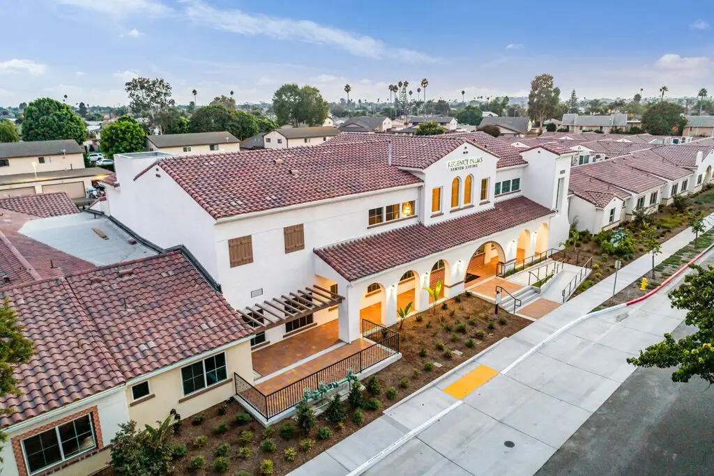Aerial view of Regency Palms Senior Living in Oxnard, a white stucco building with a red-tile roof, arched entry colonnade, and a gable sign reading “REGENCY PALMS SENIOR LIVING.” A sidewalk, planting strip, and neighboring tile-roof wings surround the campus under a blue sky.