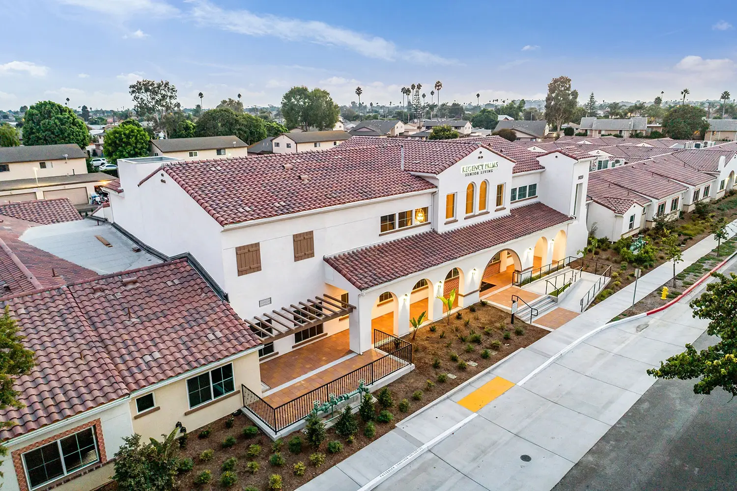 Aerial view of Regency Palms Senior Living in Oxnard, a white stucco building with a red-tile roof, arched entry colonnade, and a gable sign reading “REGENCY PALMS SENIOR LIVING.” A sidewalk, planting strip, and neighboring tile-roof wings surround the campus under a blue sky.