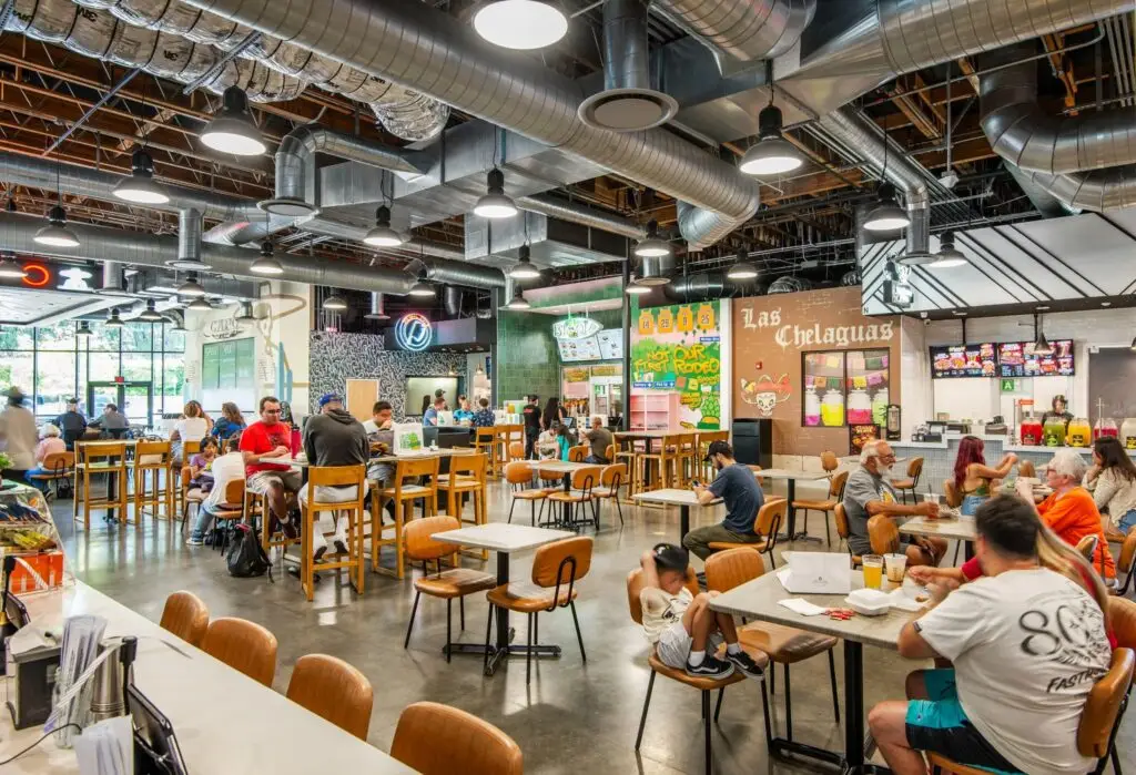 Dining hall at Rodeo 72 with shared tables, exposed ductwork, and tenant storefronts including Las Chelaguas