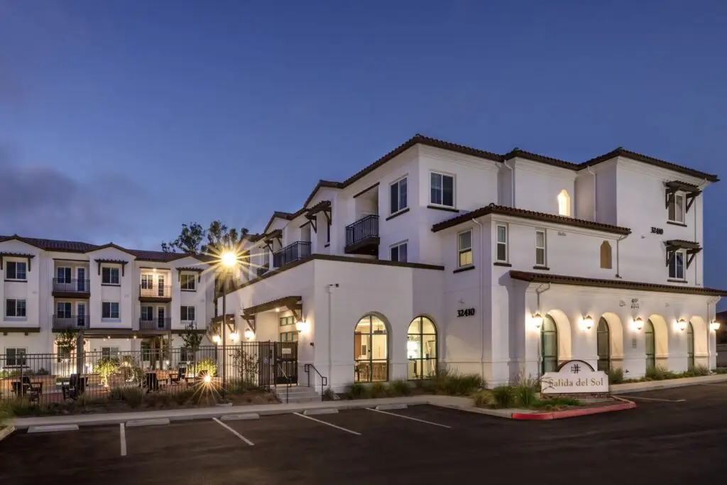 Dusk view of Salida del Sol at 32410, a three-story white stucco building with a red-tile roof, dark wood brackets, and arched ground-floor windows. A monument sign reads “Salida del Sol.” Lit amenity rooms and a fenced courtyard sit beside a parking lot under a deep blue sky.