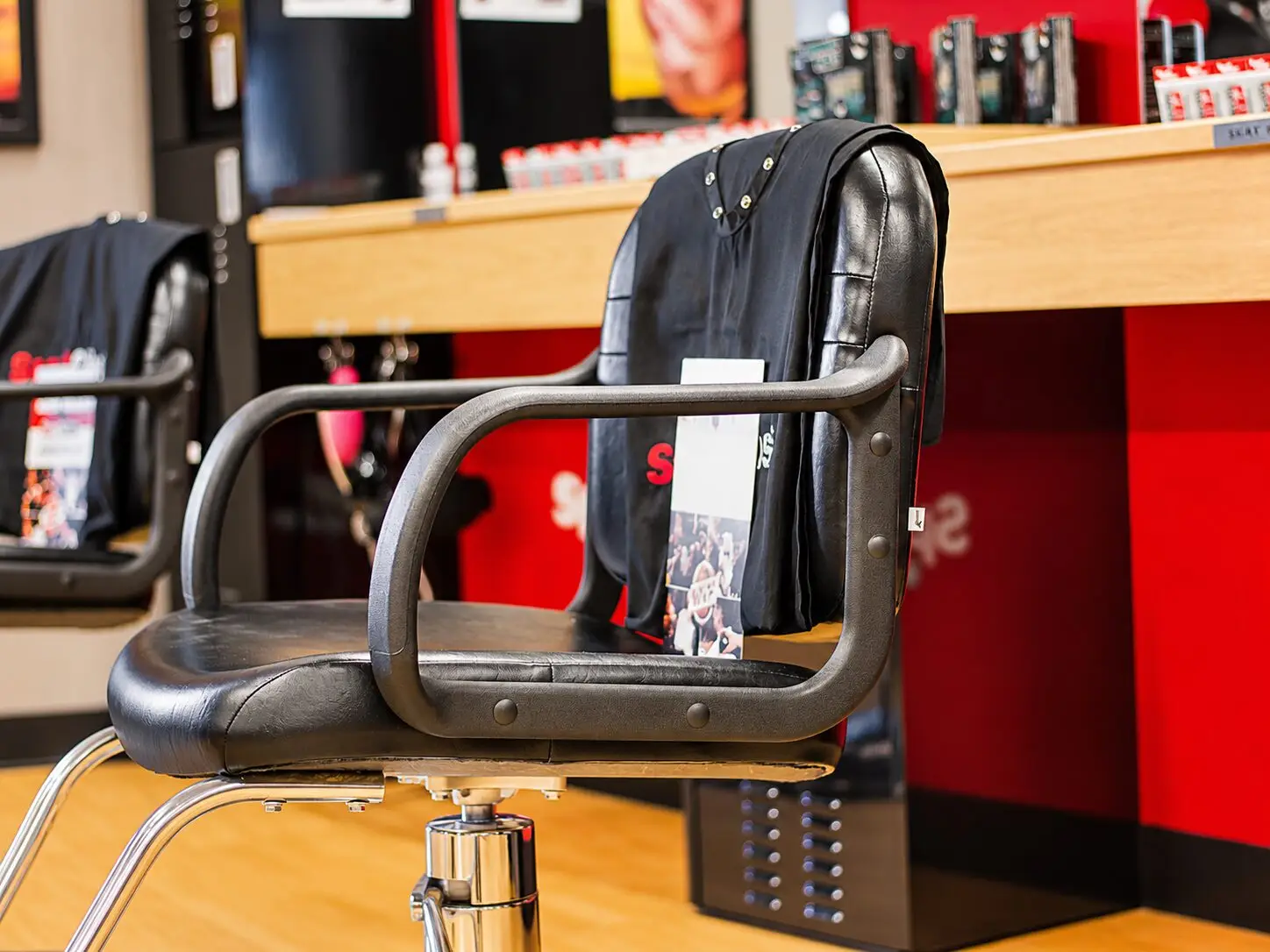 Close-up of a black leather styling chair with chrome base. A black cape and a Sport Clips magazine rest on the seat. The red station panel and wood counter are behind it.