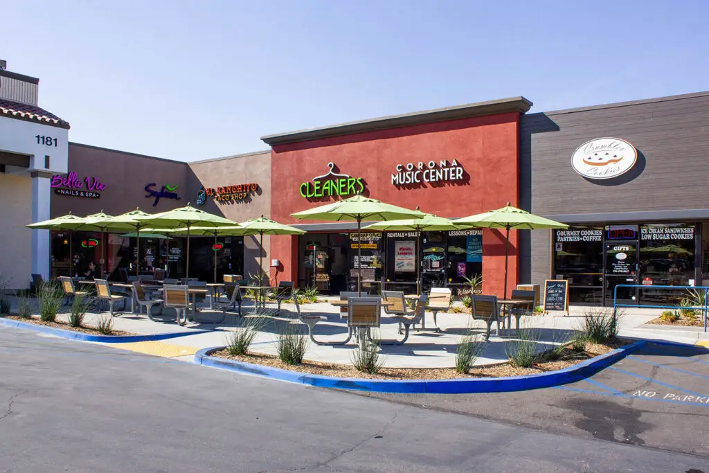 Patio in front of the red Cleaners / Corona Music Center bay, with lime umbrellas, wood tables, and neighbors including Bella Vie and Crumbl Cookies.