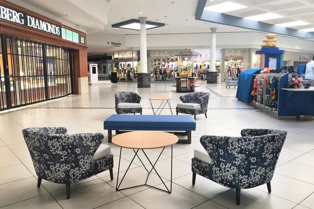 Common-area lounge at The Oaks Mall in Gainesville with floral wing chairs, a blue bench, and the corridor toward Dillard’s