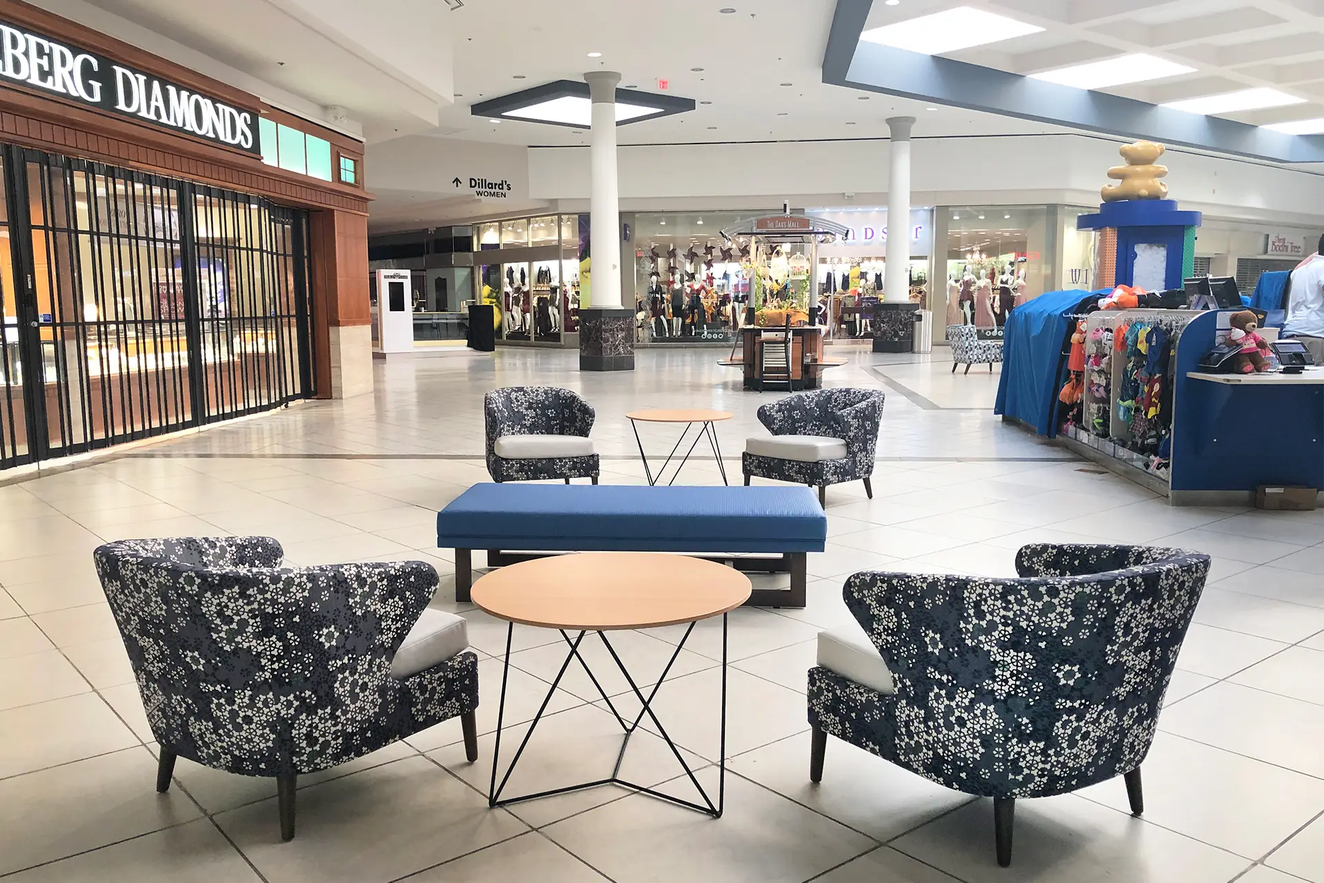 Common-area lounge at The Oaks Mall in Gainesville with floral wing chairs, a blue bench, and the corridor toward Dillard’s