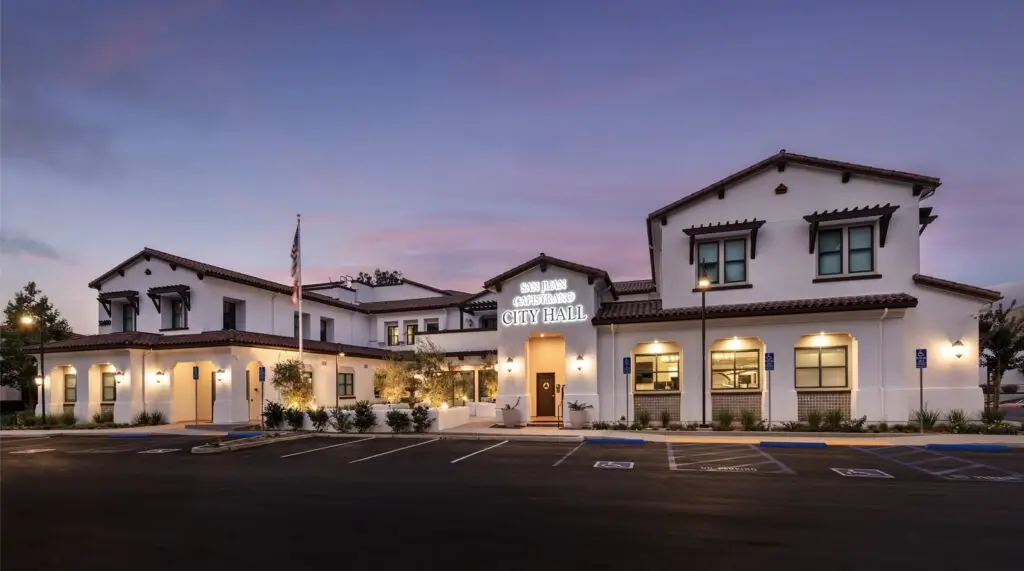 Dusk view of San Juan Capistrano City Hall with lit signage, tile roofs, and parking in the foreground