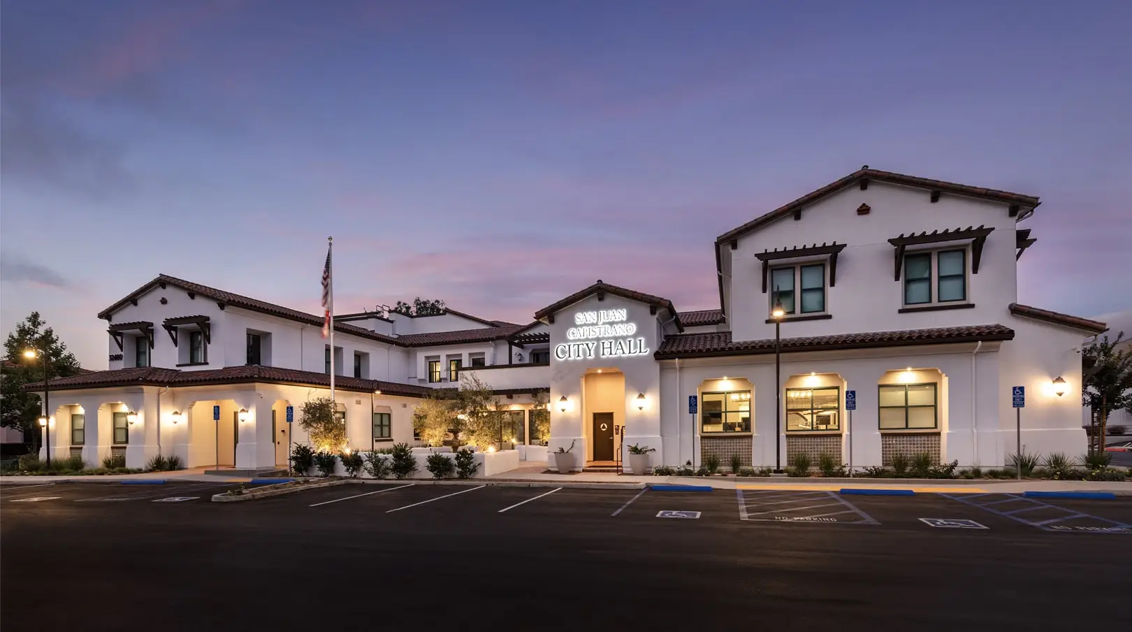 Dusk view of San Juan Capistrano City Hall with lit signage, tile roofs, and parking in the foreground