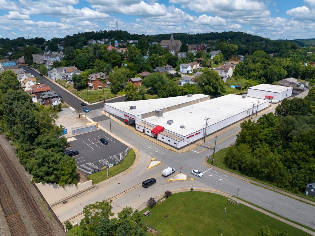 Daytime aerial of the completed OpenSpace Self Storage facility at 101 North 4th Street in Jeannette, Pennsylvania, a long white former glass-factory building with a red entry canopy, striped parking, and the railroad in the foreground.