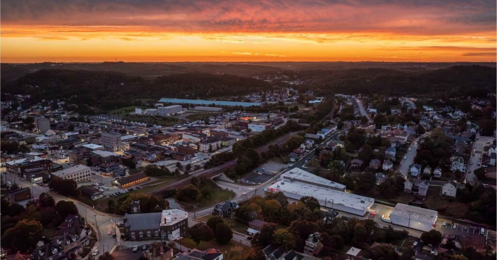 Sunset aerial over Jeannette, Pennsylvania, with the long white-roofed OpenSpace Self Storage building lit along the rail corridor, downtown blocks and church steeples in the valley, and wooded hills under an orange sky.