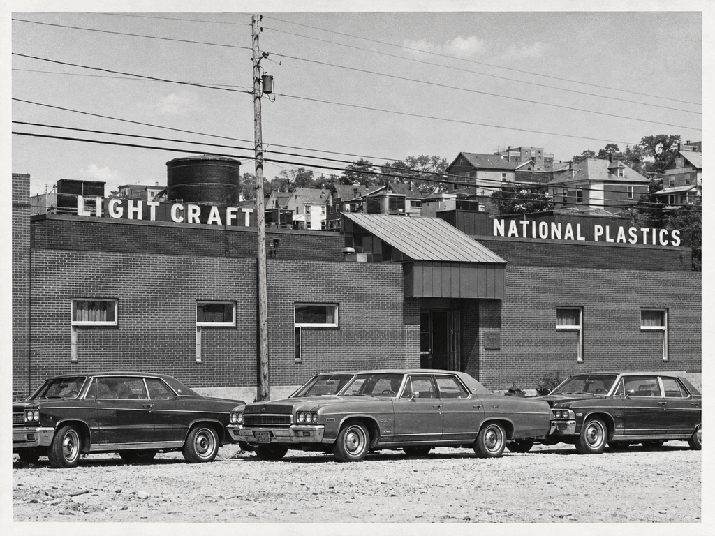 Mid-century photograph of the brick industrial building later reused for OpenSpace, with rooftop signs reading Light Craft and National Plastics, a water tower, and 1970s cars parked on the gravel lot.